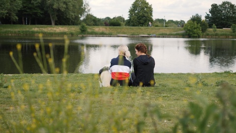 Zwei Personen sitzen mit einem Hund auf einer Wiese am Ufer eines Sees. Im Vordergrund des Bildes sind grün-gelbe Sträucher zu sehen.