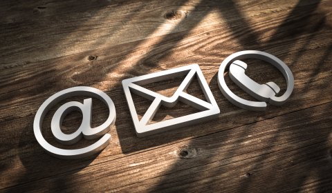 Ein @-Zeichen, ein Briefsymbol und ein Telefonhöhrer-Symbol vor einem hölzernen Hintergrund