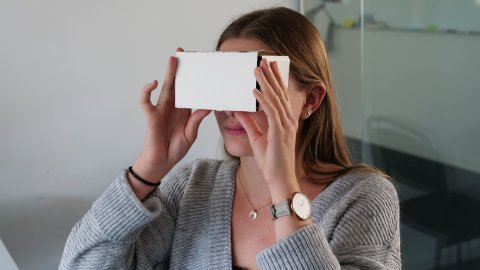 Zu sehen ist eine weibliche Person, die sich eine zusammengebaute VR-Brille aus Pappkarton vor das Gesicht hält.