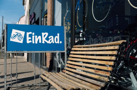 Außenansicht des Fahrradladens "EinRad". Ein blaues Schild mit dem Namen des Geschäfts ist zu sehen, außerdem ein Teil einer Holzbank, die vor dem Schaufenster steht.