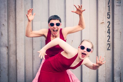 Zwei Tänzerinnen in roten Kleidern und mit rosa Sonnenbrillen bei einer Tanzperformance.
