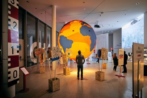 Ein Mann steht vor einer großen leuchtenden Weltkugel. Um ihn herum sind Demonstration Schilder mit klimapolitischen Sprüchen aufgestellt. 