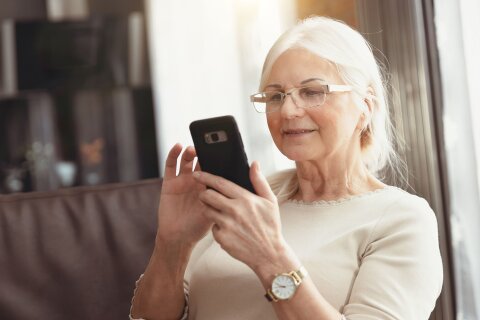 Eine ältere Frau sitzt auf einem Sofa und nutzt ein Smartphone.