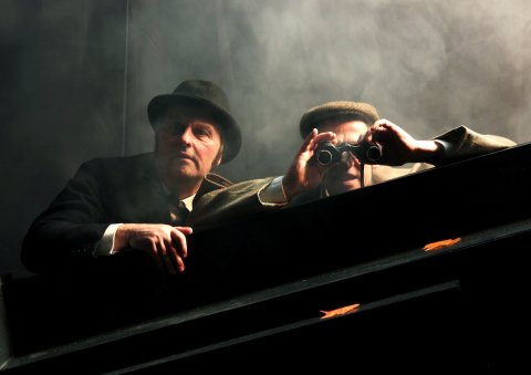 Zwei Schauspieler suchen auf der Bühne jemanden mit einem Fernglas.