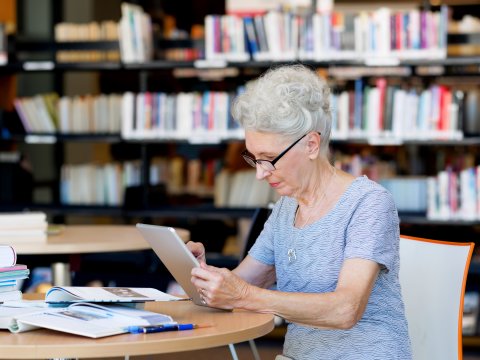 Eine ältere Dame sitzt mit ihrem iPad in der Bibliothek und studiert