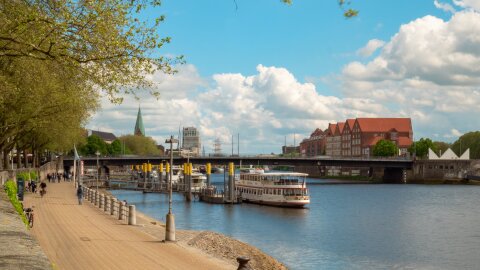 Blick die Weserpromenade entlang, am rechten Ufer ist die Weserburg zu sehen, auf dem Wasser einige Schiffe, am linken Ufer Passantinnen und Passanten sowie grün bewachsene Bäume.