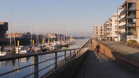 Links im Bild ist ein kleiner Yachthafen mit Booten zu sehen. Rechts davon verläuft eine Promenade, die mit Häusern bebaut ist. Radfahrer fahren die Promenade entlang.