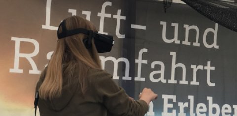 Ein Mädchen hat eine VR-Brille auf. Hinter ihr stehen die Worte "Luft- und Raumfahrt erleben"