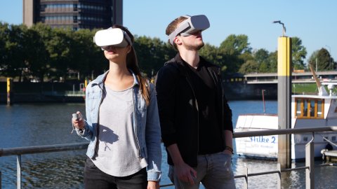 Zu sehen sind zwei Personen, die eine große Brille tragen. Sie stehen an einem Geländer, hinter dem Wasser zu sehen ist. Rechts im Bild ist ein Teil eines Schiffes zu erkennen; im Hintergrund stehen Bäume und ein braunes Gebäude.