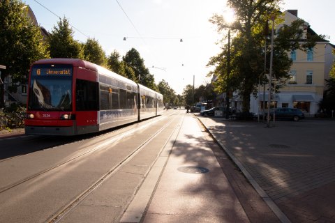 Blick in die Wachmannstraße. Eine Straßenbahn fährt vorbei. Im Hintergrund sind Autos und Personen, die die Fahrbahn überqueren. Außerdem ist eine Person auf einem Dreirad zu sehen.