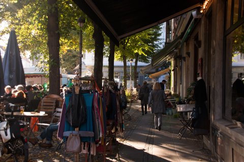 Kleidung hängt an einer Stange vor einem Geschäft, Menschen sitzen in einem Café in der Sonne