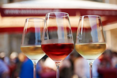 Zwei Weingläser mit einem roten und einem weißen Wein gefüllt stehen in einer Nahaufnahme vor dem Festgetümmel im Hintergrund auf einem Tisch.