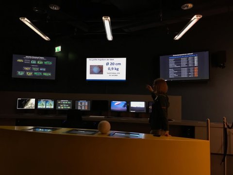 Eine Frau steht vor einem Modell eines großen Hagelkorns und mehreren Monitoren