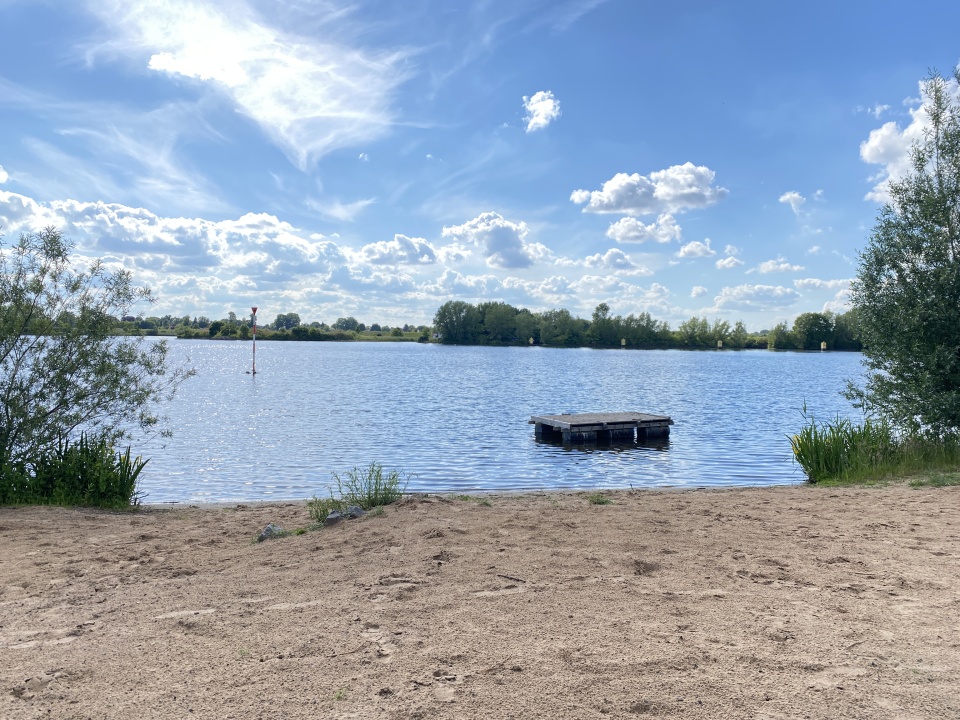An einem kleinen Sandstrand führt eine Rutsche ins Wasser. Im Hintergrund schwimmt eine Palette in einem See.