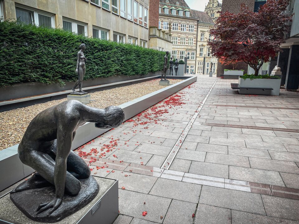 Mehrere menschliche Figuren stehen verteilt in einem langen Gang. Bäume verzieren den Weg.