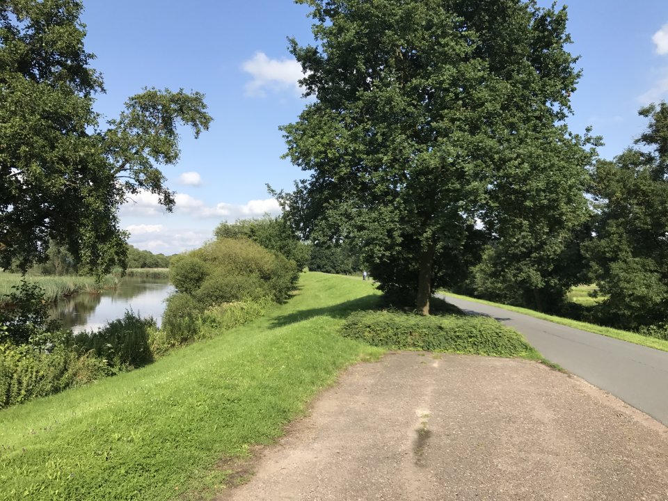 Rechts ist ein Weg auf einem Deich zu erkennen. Rechts im Bild ist ein Fluss zu sehen, der von grünen Bäumen und Wiesen umgeben ist.