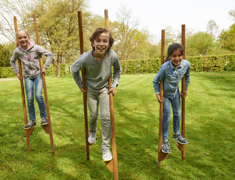 Zu sehen sind drei Kinder, die mit hölzernen Stelzen auf einer grünen Rasenfläche laufen.