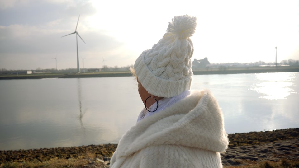 Zu sehen ist eine Person von hinten, die eine weiße Jacke und eine weiße Mütze trägt und am Ufer steht. Von dort aus sieht man das Wasser der Weser und das gegenüberliegende Ufer, auf dem Häuser und Windräder stehen.