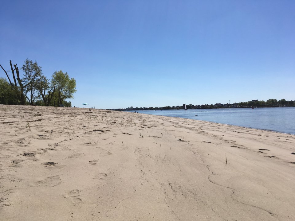 Zu sehen ist ein langer, weißer Sandstrand, an dessen rechter Seite blaues Wasser grenzt. Links vom Strand stehen grüne Bäume. Der Himmel ist strahlend blau.