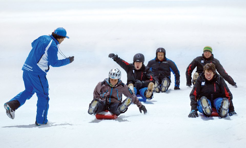Fünf Personen rodeln im Schnee um die Wette, eine sechste nimmt die Zeit.