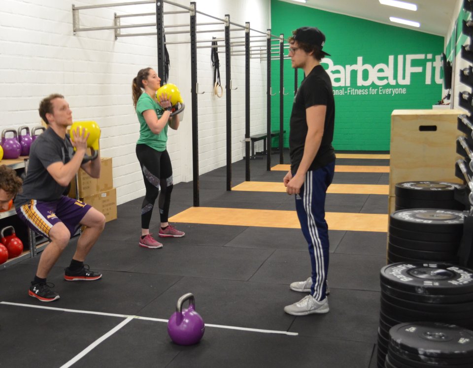 Ein Mann und eine Frau trainieren im Fitnessstudio mit Gewichten. Ein anderer Mann steht vor ihnen und schaut, ob die beiden die Übungen richtig ausführen. 