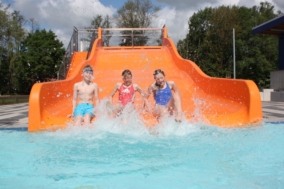 Zwei Mädchen und ein Junge sitzen in ihren Badesachen auf einer orangenen Wasserrutsche im Freien.