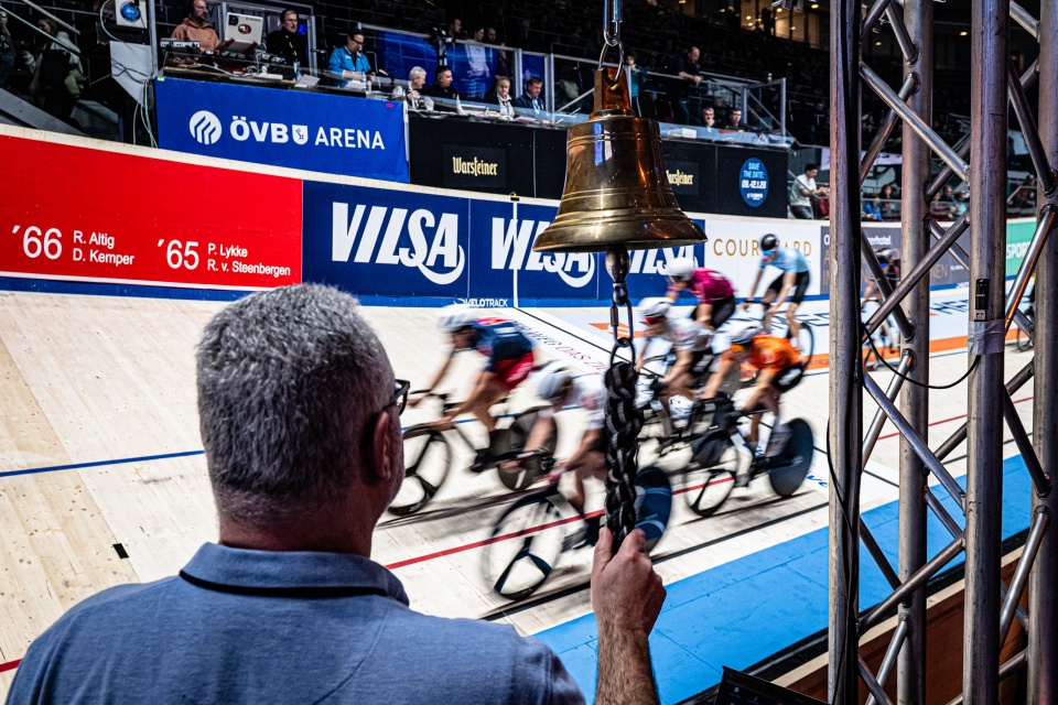 Männer fahren auf Rädern ein Rennen aus. Ein Mann steht an einer Glocke.