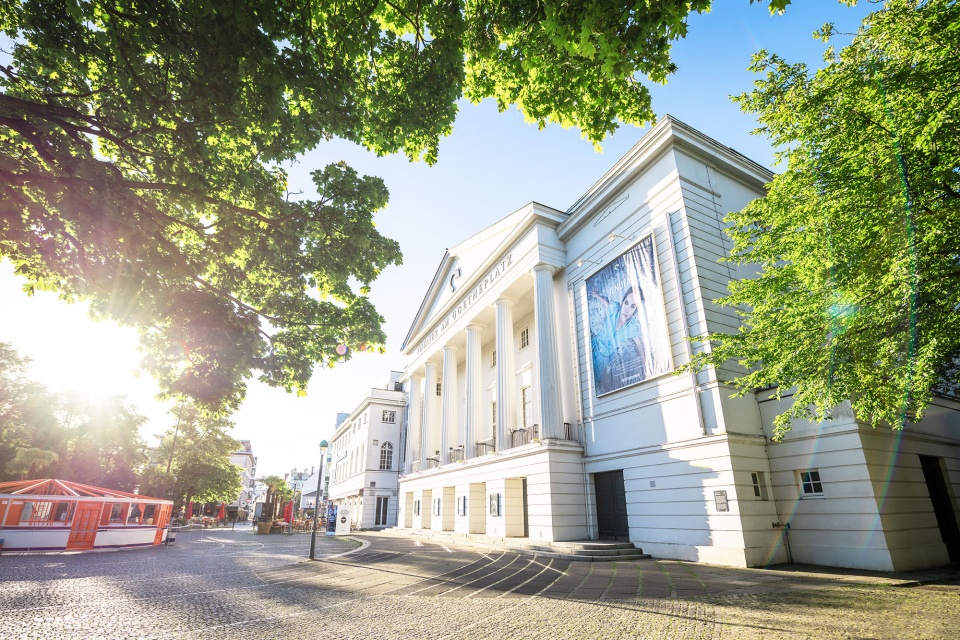 Blick durch grüne Bäume auf das Theater am Goetheplatz an einem sonnigen Tag. 