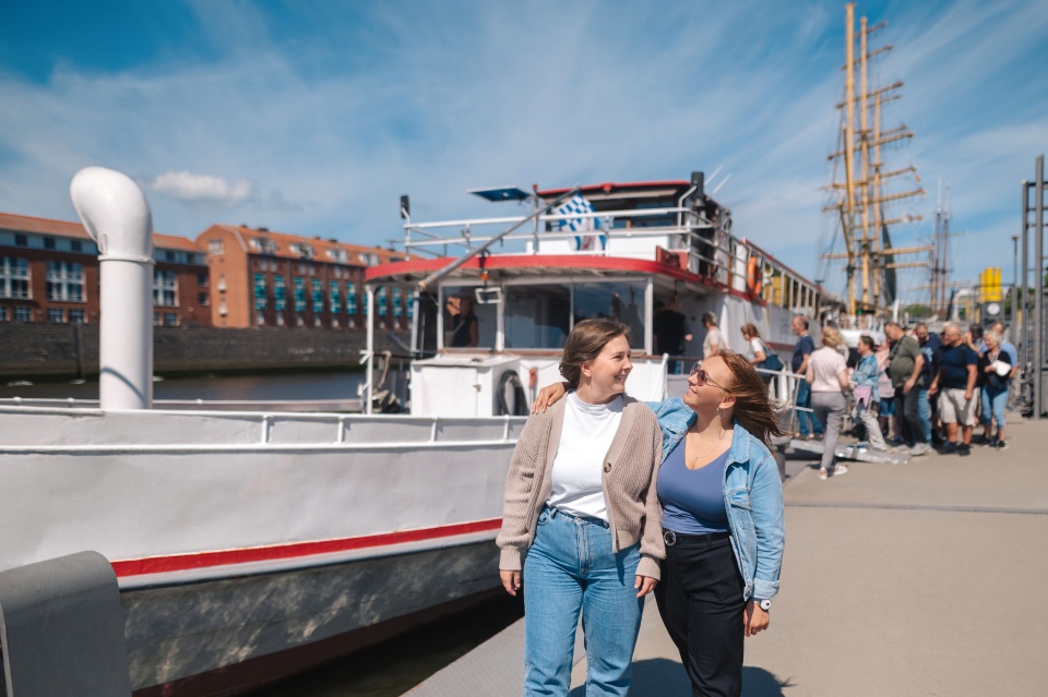 Two women laugh at each other. In the background, people are boarding a ship.