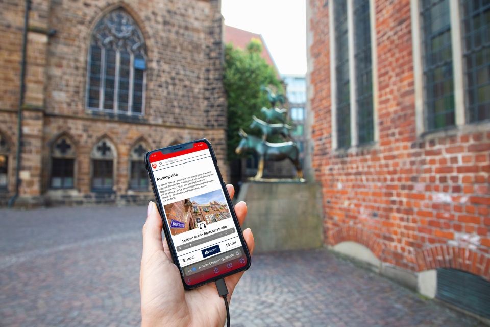 Jemand hält ein Smartphone, auf dem der Audioguide geöffnet ist. Im Hintergrund sind die Bremer Stadtmusikanten.