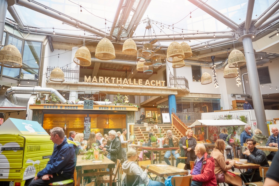 Innenansicht der Markthalle 8, einer Halle in der viele verschiedene Restaurants und Tische sind, an denen Menschen sitzen.