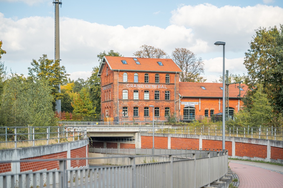 Eine Straße führt unter Gleisen lang. Im Hintergrund ist ein altes Bahnhofsgebäude zu sehen.