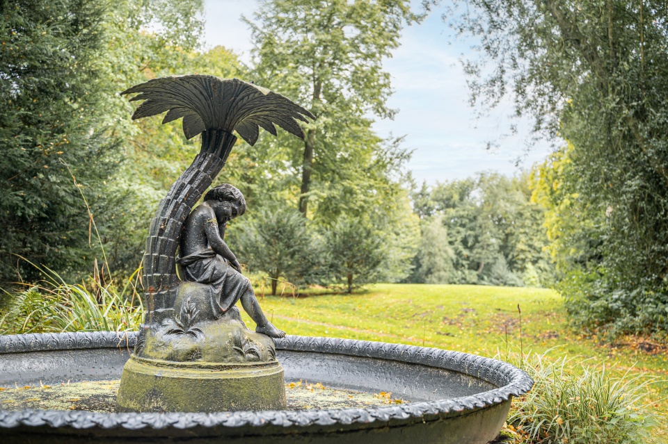 Schwarzer Brunnen im Grünen in dessen Mitte eine Insel ist, auf welcher ein Kind sitzt und sich ein Stein und eine Palme befinden. 