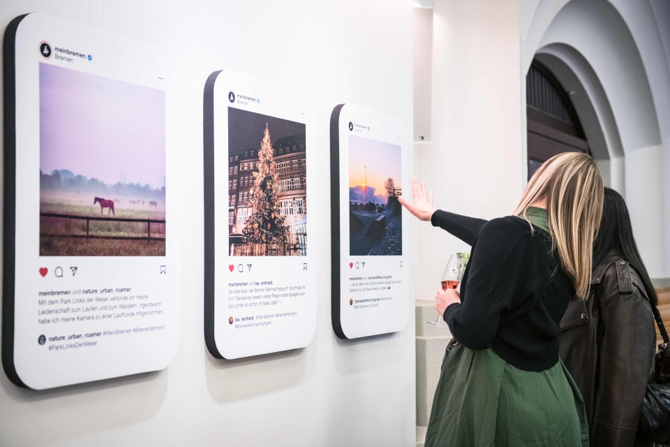 An einer weißen Wand hängen drei Fotos, die Bremer Motive zeigen. Davor stehen zwei Personen, die diese Fotos betrachten.