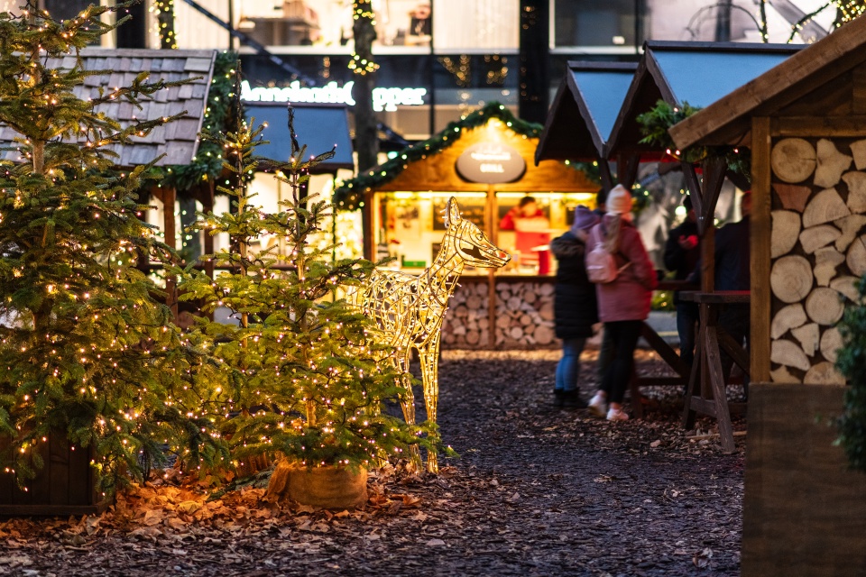 Tannenbäume und beleuchtete Buden auf einem Wintermarkt, Menschen stehen an einem überdachten Tisch.