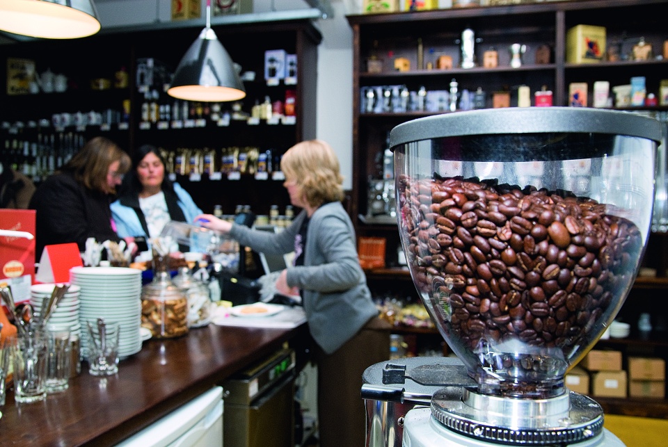 Eine Mitarbeiterin bedient im Lloyd Caffee zwei Kundinnen. Im Vordergrund sind Kaffeebohnen in einer Maschine zu sehen. 