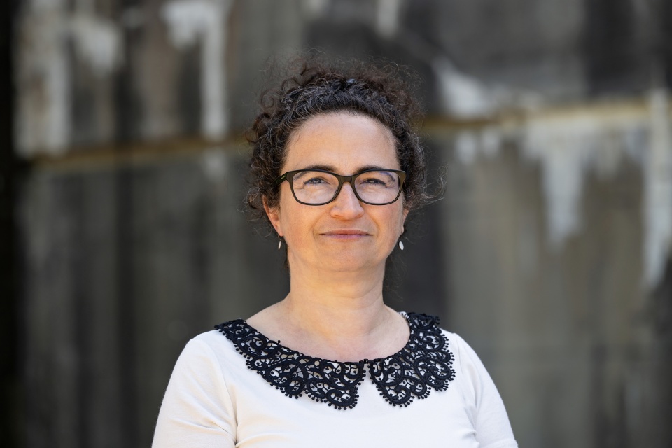 Eine Frau in einem weißen Tshirt, schwarzer Brille und hochgebundenen schwarzen Haaren lacht. 