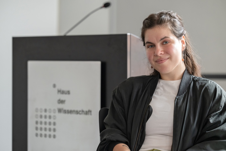Porträtfoto einer Frau mit braunen Haaren. Hinter ihr steht auf einem Rednerpult: "Haus der Wissenschaft".