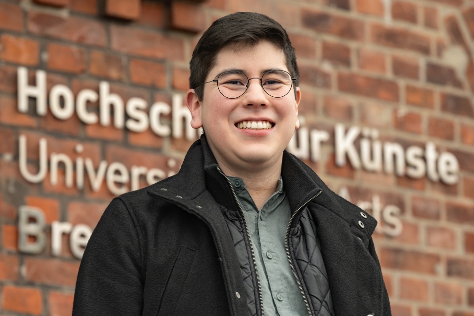 Ein Wissenschaftler schaut lächelnd in die Kamera. Hinter ihm befindet sich das Gebäude von der Hochschule für Künste.