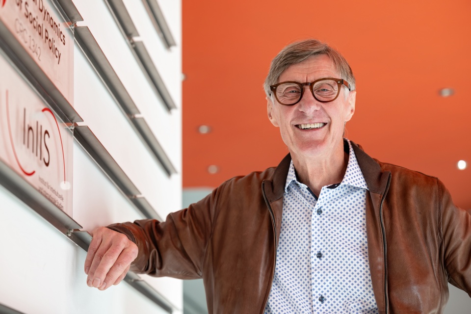 Ein Mann mit Brille steht vor einem orangenem Hintergrund und lacht