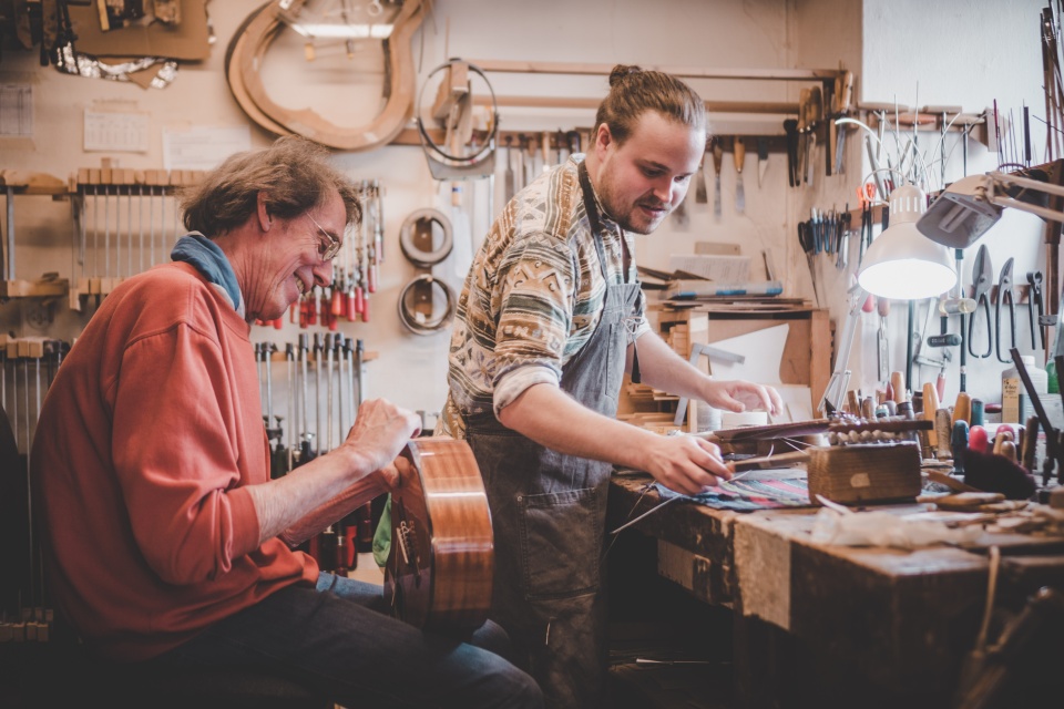 Ein älterer und jüngerer Mann arbeiten gemeinsam an der Werkbank an einer Gitarre.