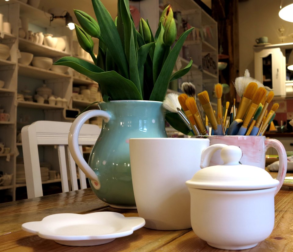 Mehrere Keramikgefässe, die auf einem Holztisch stehen. In eine Keramiktasse wurden Pinsel gesteckt, in einer weiteren grüne Pflanzen.Im Hintergrund sind Regale mit weiteren Keramikprodukten zu sehen. 
