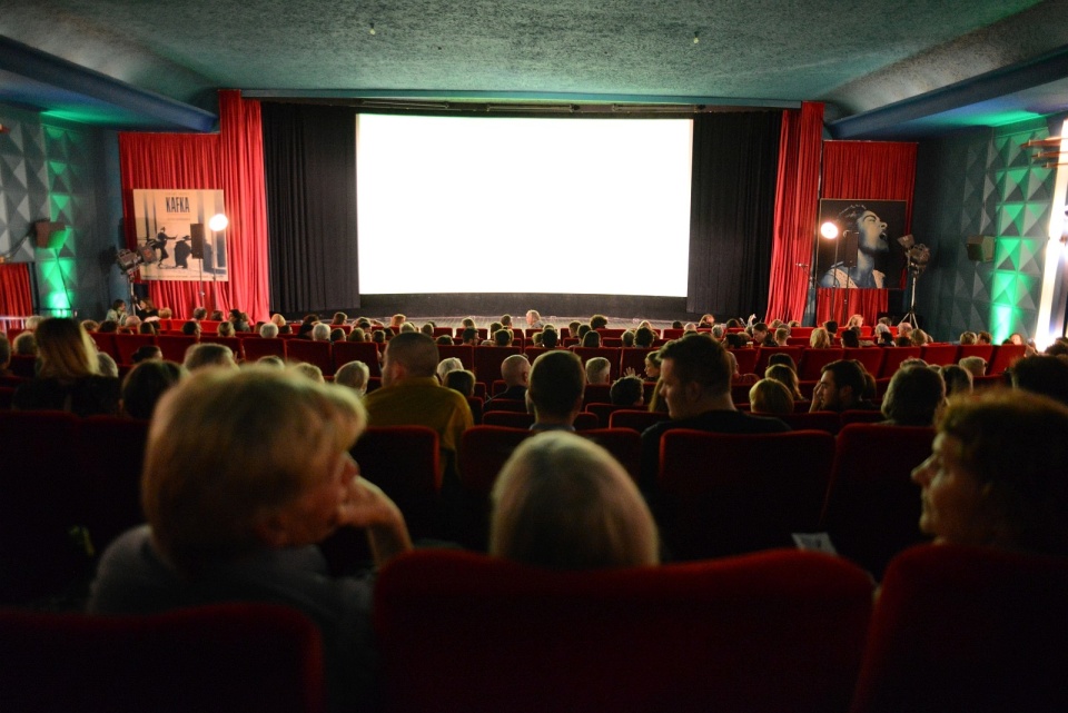 Viele Menschen sitzen in einem Kino. Vorne ist die Leinwand zu sehen. Links und rechts sind schwarze und rote Vorhänge. 