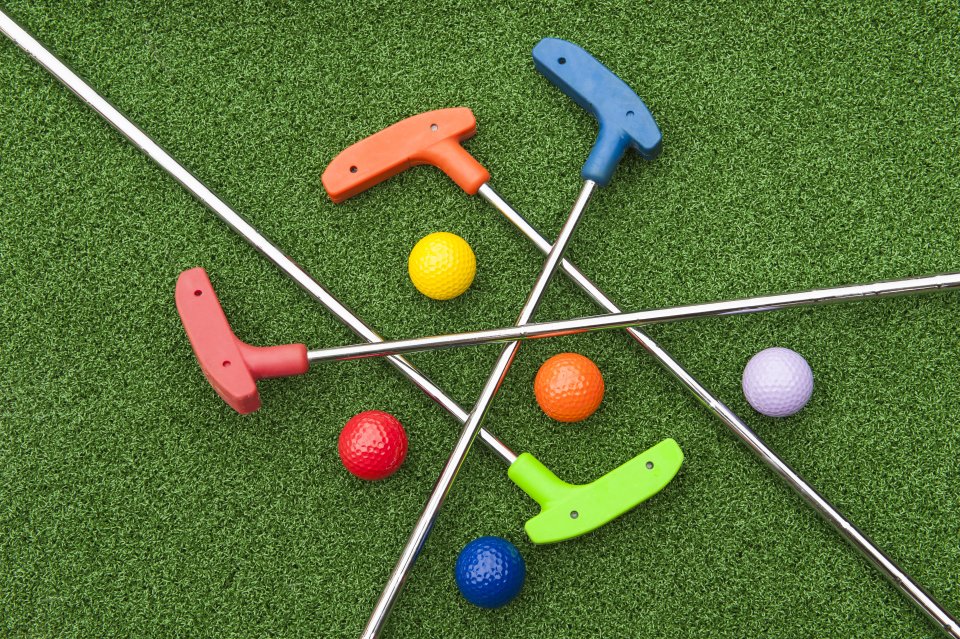 Zu sehen sind vier Minigolfschläger in verschiedenen Farben und passende Bälle auf einer Grünfläche.