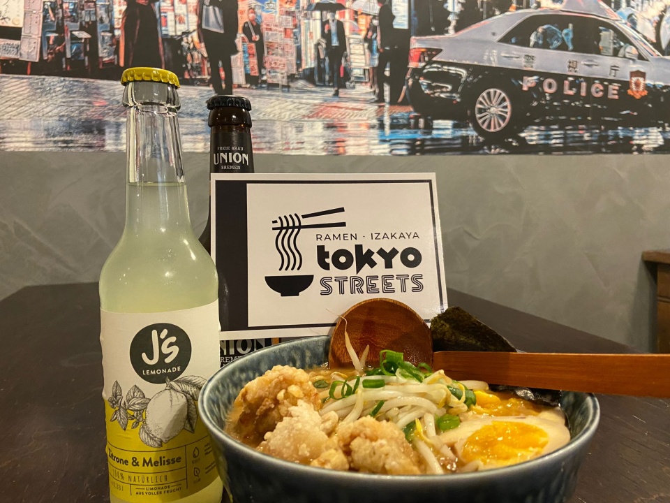 Ein gedeckter Tisch mit einer Schüssel Ramen, einer Limonade, einem Bier und einer Gutscheinkarte von Tokyo Streets