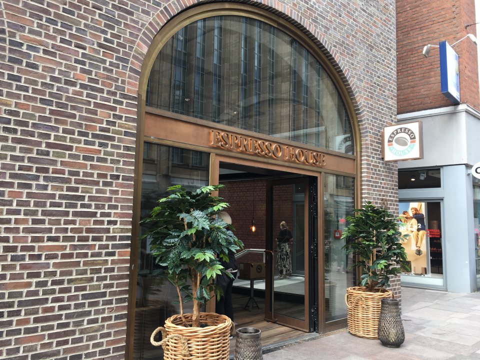 Das Espresso House Bremen in der Außenansicht. Vor der Tür stehen zwei kleine Bäume in geflochtenen Körben. Die Tür ist groß. Über der Tür steht "Espresso House". 
