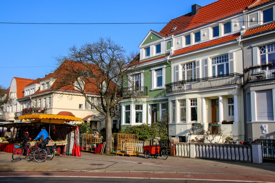 Vor drei bunten alten Häusern stehen zwei Wochenmarktstände und ein Mann hält dort mit einem Fahrrad. 