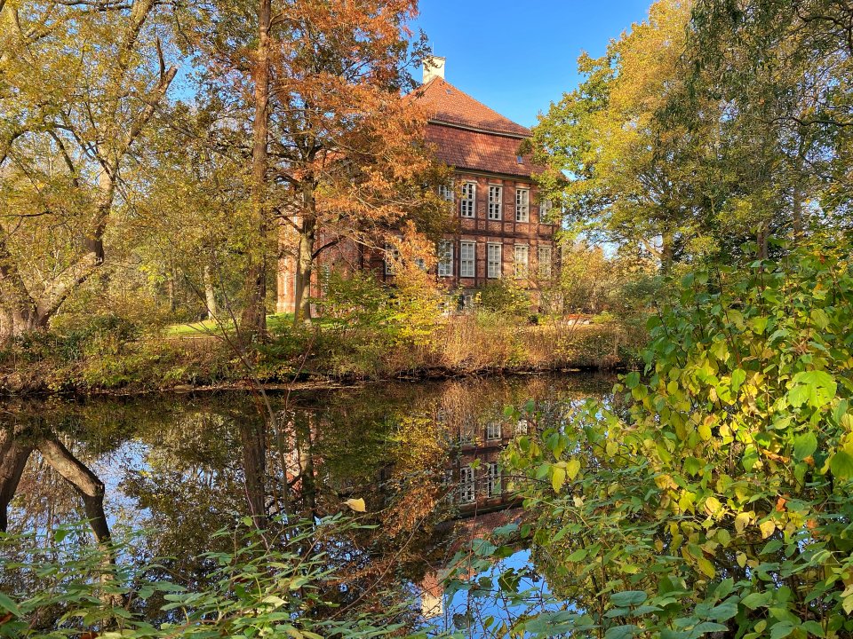 Das von herbstlichen Bäumen und Büschen sowie einem Gewässer umgebene Schloss Schönebeck an einem sonnigen Herbsttag.