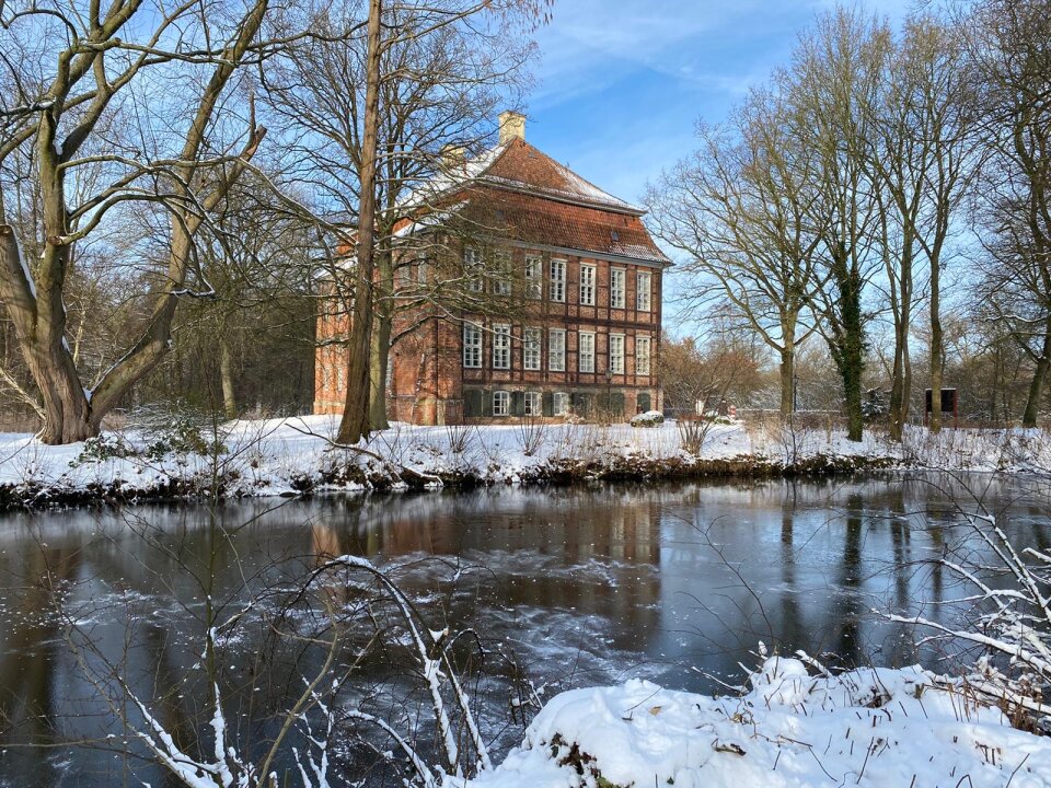 Ein altes Gebäude steht in einem Park. Davor fließt ein kleiner, zugefrorener Fluss. 