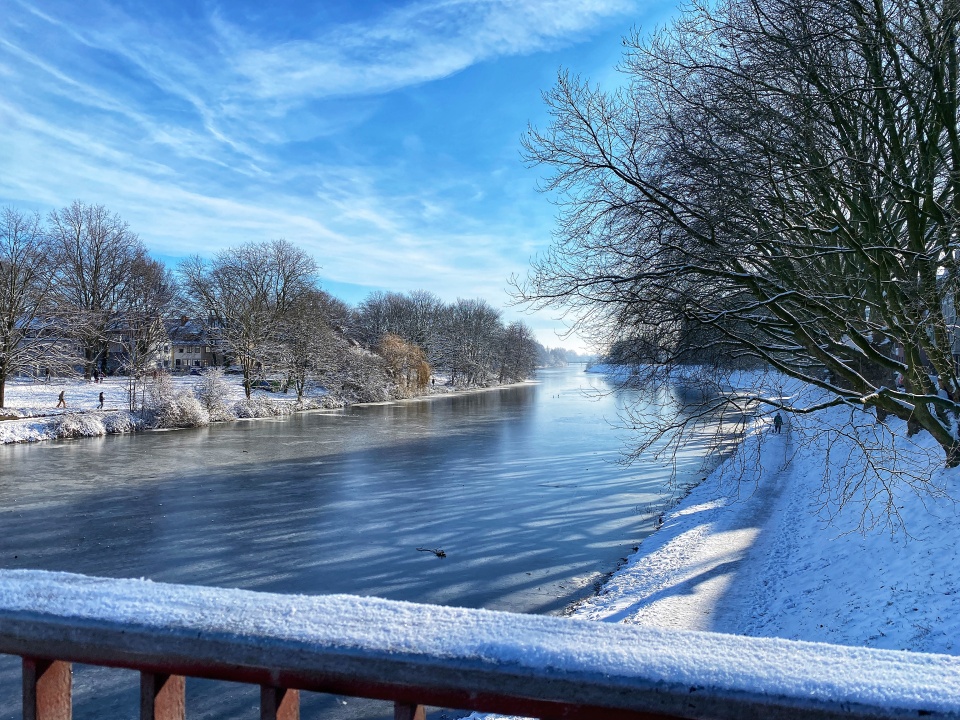 Der Blick geht über ein Gelände auf den Werdersee. Er ist zugefroren. Die Natur am Rand ist mit Schnee bedeckt und man sieht Leute spazieren.